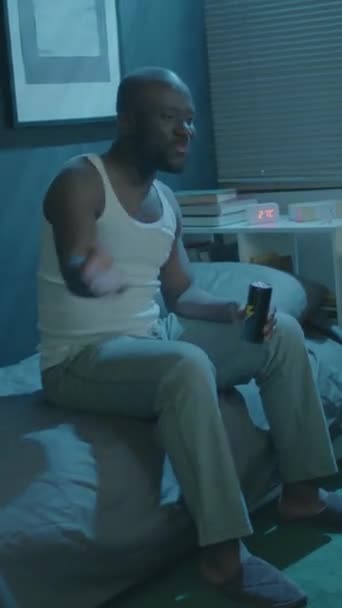 黑人男子坐在床上 喝能量饮料 支持运动队 同时在晚上看电视比赛的垂直镜头 — 图库视频影像