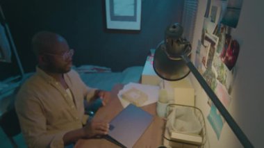 Afro-Amerikalı adamın dizüstü bilgisayarını kapatırken, masa lambasını kapatırken ve gece geç saatlere kadar çalışırken yatarken yüksek açılı görüntüsü.