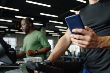 Bilinmeyen beyaz adam spor salonunda eğitim görüyor elinde akıllı telefon, yaşlı siyah adam arka planda egzersiz yapıyor.