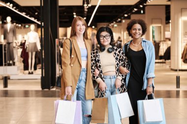 Alışveriş merkezinde kıyafet dükkanına karşı poz veren çok ırklı bir grup kadın kameraya bakıp gülümsüyor.