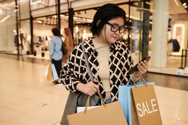 Young asian shopping mall visitor holding bags with bought items while checking her mobile phone