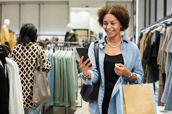 Laughing young african american woman looking at smartphone screen while shopping at clothing store
