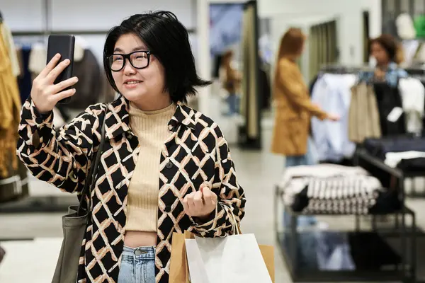Young asian fashion blogger filming herself on smartphone walking through clothes store at mall