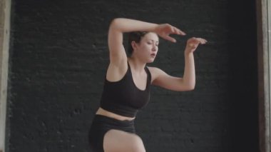 Siyah dans stüdyosunun arka planında akrobatik unsurlarla zarif çağdaş dans eden genç ve esnek bir kadının çekimini indirin.