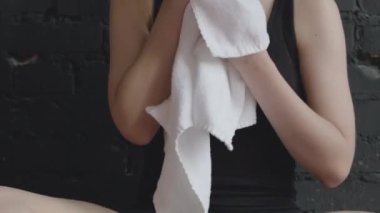 Beyaz havluyla terini ve yüzünü silerken spor salonundaki siyah tuğla duvarın önünde otururken beyaz havlu ve su içerken çekilen beyaz kadın.