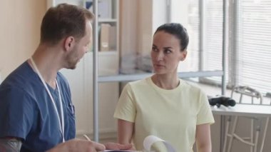 Profesyonel fizyoterapistin klinikteki randevusu sırasında panoya not aldığı ve kadın hastayla konuştuğu orta boy bir fotoğraf.