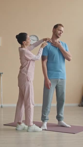 診療所の運動マットの上に立っている男性患者の腕筋を点検する女性の理学療法士の縦のショット — ストック動画