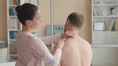 Kadın fizyoterapist, klinikte kontrol sırasında kas problemleri teşhis ederken gömleksiz bir erkeğin omuz ve koluna dokunuyor.