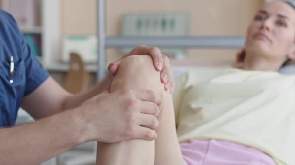 怪我後に診療所を訪れる女性患者の膝を調べるプロの理学療法士の選択的な焦点ショットを閉じる — ストック動画