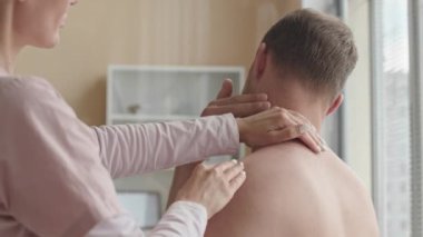 Kadın fizyoterapist, klinikteki tıbbi muayenede ağrıyan kasları teşhis ederken gömleksiz bir erkeğin omuz ve boynuna dokunuyor.