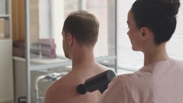 健康診療所で働いている間シャツなしの男性患者の背部筋にマッサージ銃を使用して専門の女性の理学療法士 — ストック動画