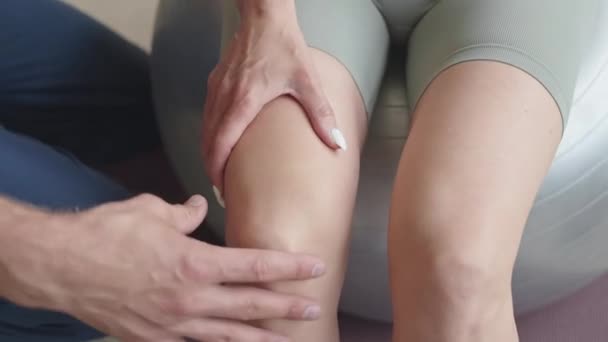 リハビリテーション療法中にフィットネスボールに座っている女性患者の膝関節と関節をチェックする理学療法士の手の近いショット — ストック動画