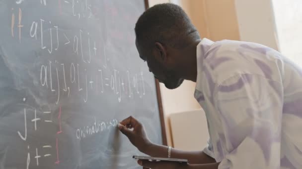 デジタルタブレットを保持し 教室でチョークボード上のコンピュータアルゴリズムを書く若いアフリカ系アメリカ人の大学生 — ストック動画