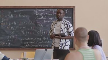 Genç Afrikalı Amerikalı öğretmen elinde tablet tutuyordu ve üniversitedeki programlama dersleri sırasında çeşitli öğrencilerle konuşuyordu.