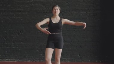 Beyaz kadın balerin siyah aktivite giysileri giyerken orta ölçekli bir fotoğraf. Çatı katı tarzı bale stüdyosunda siyah tuğla duvara karşı duran koreografi dersi veriyor.