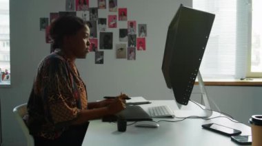 Stüdyoda bilgisayar başında çalışırken stilus kalem ve resim tableti kullanan Afro-Amerikalı kadın grafik tasarımcısı.