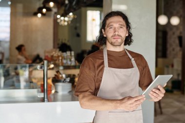 Yemek yiyen erkek çalışan, iş yerinde kameraya bakan tabletle poz veriyor.