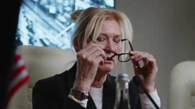 Üst düzey kadın politikacı mikrofonla konuşuyor ve askeri basın toplantısı sırasında gözlüklerini çıkarıyor.