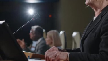 Üst düzey kadın politikacı mikrofonla podyuma çıkıyor ve basın toplantısı sırasında seyircilerin önünde askeri durum hakkında rapor veriyor.