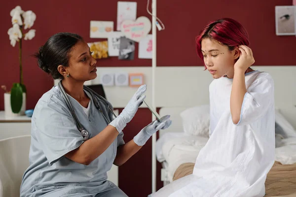 Young plastic surgeon holding mirror in front of teenage patient while consulting her before cosmetic operation or procedure