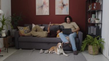 Küçük bir av köpeğiyle evdeki kanepede dinlenen ırklar arası bir çiftin uzun bir çekimi. Çift ırklı kız, beyaz erkek arkadaşının kalçasında dijital tabletle yatarken, o dizüstü bilgisayarda geziniyor.