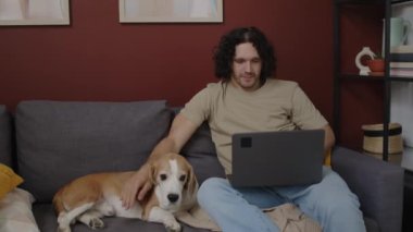 Orta boylu, uzun kıvırcık saçlı, yakışıklı beyaz bir adamın şirin köpeğini okşayıp dizüstü bilgisayarında gezerken rahat bir şekilde şık bir dairede kanepede oturduğu bir fotoğraf.