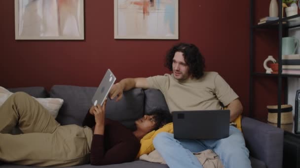 在他们的公寓里 一对恋爱中的跨种族年轻夫妇躺在沙发上 带着数码平板电脑和笔记本电脑 一个骨瘦如柴的女人躺在她的白人男朋友身旁 一边聊天一边笑 — 图库视频影像