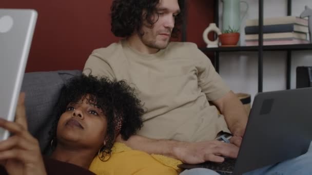 中午时分 美丽的年轻美女和她英俊的白种人男友一起躺在沙发上 在数码平板电脑和笔记本电脑上浏览着 拍了一张中等的照片 — 图库视频影像