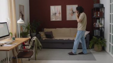 Evden çalışan, modern oturma odasında ileri geri yürüyen, akıllı telefondan konuşan, günlük kıyafetli genç beyaz iş adamlarının tam karesi.