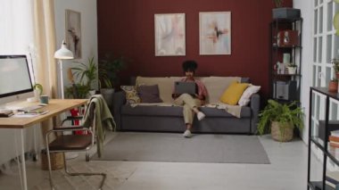 Laptop kullanan, cep telefonuyla konuşan, modern oturma odasındaki gri koltukta oturan ve evden çalışan genç, çekici, çift ırklı bir iş kadını.