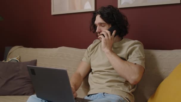 快適なホームオフィスでソファーに座っているラップトップで働いている間 スマートフォンで話す白人若いビジネスマンのミディアムショット — ストック動画