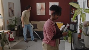 Irklar arası genç çiftin modern bir dairede birlikte ev işi yaparken, iki ırklı bir kadının mikrofiber bez ile yüzeylerin tozunu alırken ve beyaz bir adamın süpürme yaparken orta boy fotoğrafı.