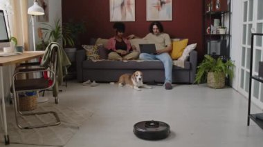 Köpeği olan ırklar arası genç çift dijital tablet ve dizüstü bilgisayar kullanırken aynı zamanda lüks oturma odasındaki kanepede dinlenirken robot elektrikli süpürge çalışıyor.