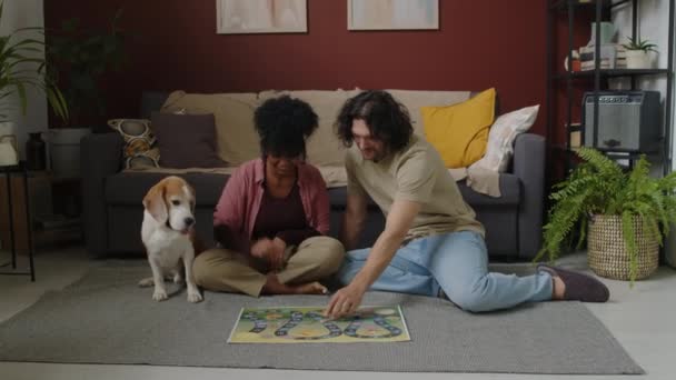 在现代客厅里 一对年轻的跨种族夫妇和可爱的小猎犬一起坐在地板上聊天和玩木板游戏 — 图库视频影像