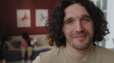 Orta boy beyaz adam portresi, uzun kıvırcık siyah saçlı, kameraya gülümseyen, şık oturma odasında duran, melez bir kadın, bulanık arka planda, kanepede köpekle oynarken.