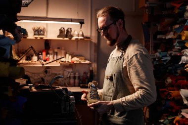 Ayakkabı yapan genç sakallı ayakkabıcının yan görünüşü. Ayakkabı yapımı sırasında elinde çizmelerle dikiş makinesinin önünde duruyor.