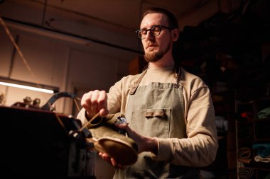 Genç erkek üretici dikiş makinesinde iplik gererken yeni ayakkabılar üretmek için elektrikli ekipman kullanıyor.