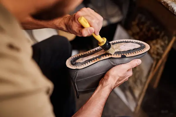 Hand of unrecognizable male shoemaker spreading glue over leather sole of grey boot while sitting in workshop and creating new footwear