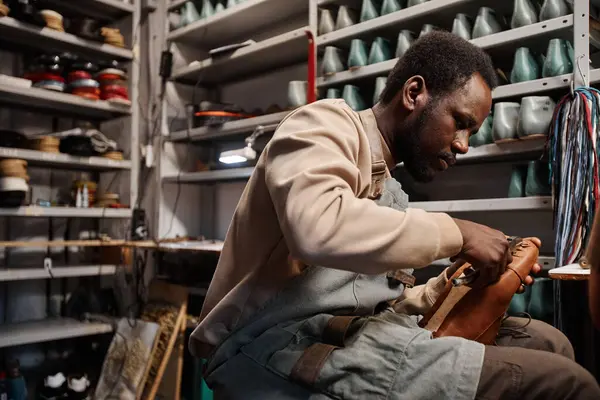 Stock image Young African American male owner of shoemaking workshop fixing upper part of unfinished boot to sole while sitting by workplace