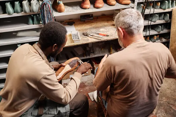 Stock image Young African American apprentice of shoemaker fixing upper part of boot to sole while sitting next to his master and repeating after him