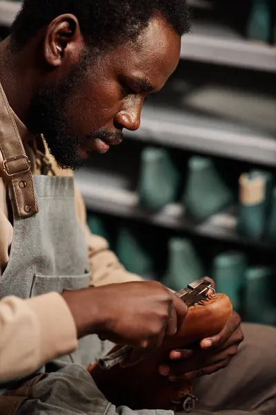Side view of young African American man in apron creating new boot or shoe in workshop and fixing together upper part and sole