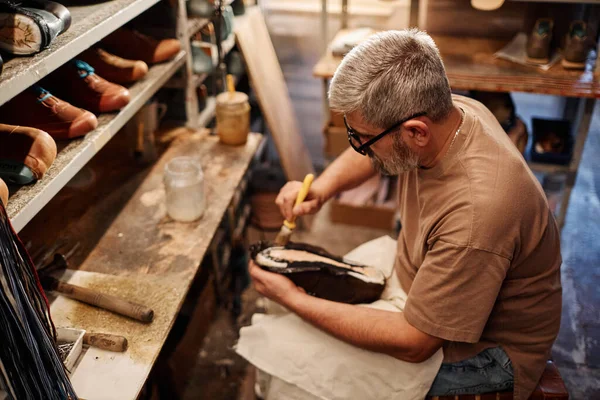 Side view of mature experienced shoemaker spreading glue over sole of unfinished boot or shoe while sitting by his workplace