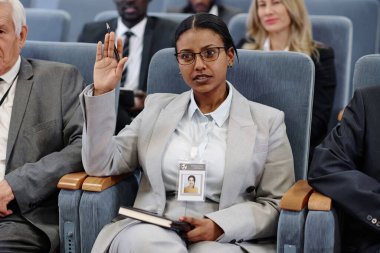 Takım elbiseli genç iş kadını iş konferansında otururken elini kaldırıyor.