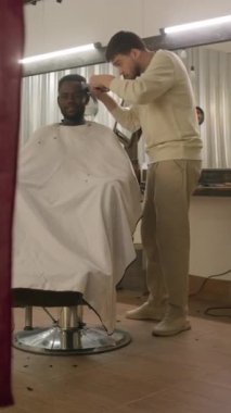 Kuaför pelerinli genç Afrikalı Amerikalı müşterinin kuaförde profesyonel berber tarafından yeni saç kesiminin dikey çekimi.