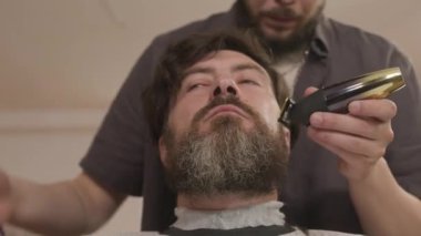 Berberin, saç makasıyla erkek müşterinin sakalını budarken ve tarak ile tararken düşük açılı görüntüsü.