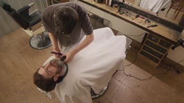 Kuaför pelerinli bir adamın kuaförde arkasına yaslandığı ve profesyonel berberin sakalını budayarak tıraş ettiği yüksek açılı görüntü.