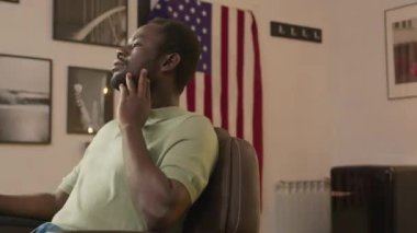 Berber dükkanında Amerikan bayrağının önünde oturmuş gülümseyerek kameraya poz veren neşeli Afro-Amerikan genç adamın portresi.