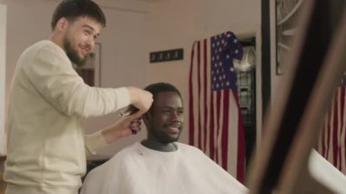 Berber neşeli Afrikalı Amerikalı müşteriyle sohbet ediyor ve saçını saç makasıyla kesiyor.