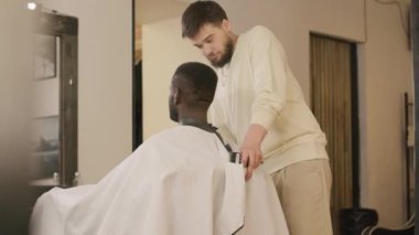 Kafkasyalı berber, berber dükkanında sakalını tımar etmeden önce Afrikalı Amerikalı erkek müşteriye sandalyeye yaslanmasında yardım ediyor.