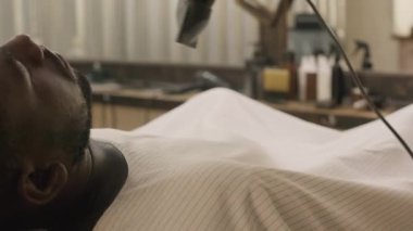 Kuaför pelerinli siyah adam gözleri kapalı otururken berber saç kurutma makinesi ve fırçayla gevşek saçlarını temizliyor.